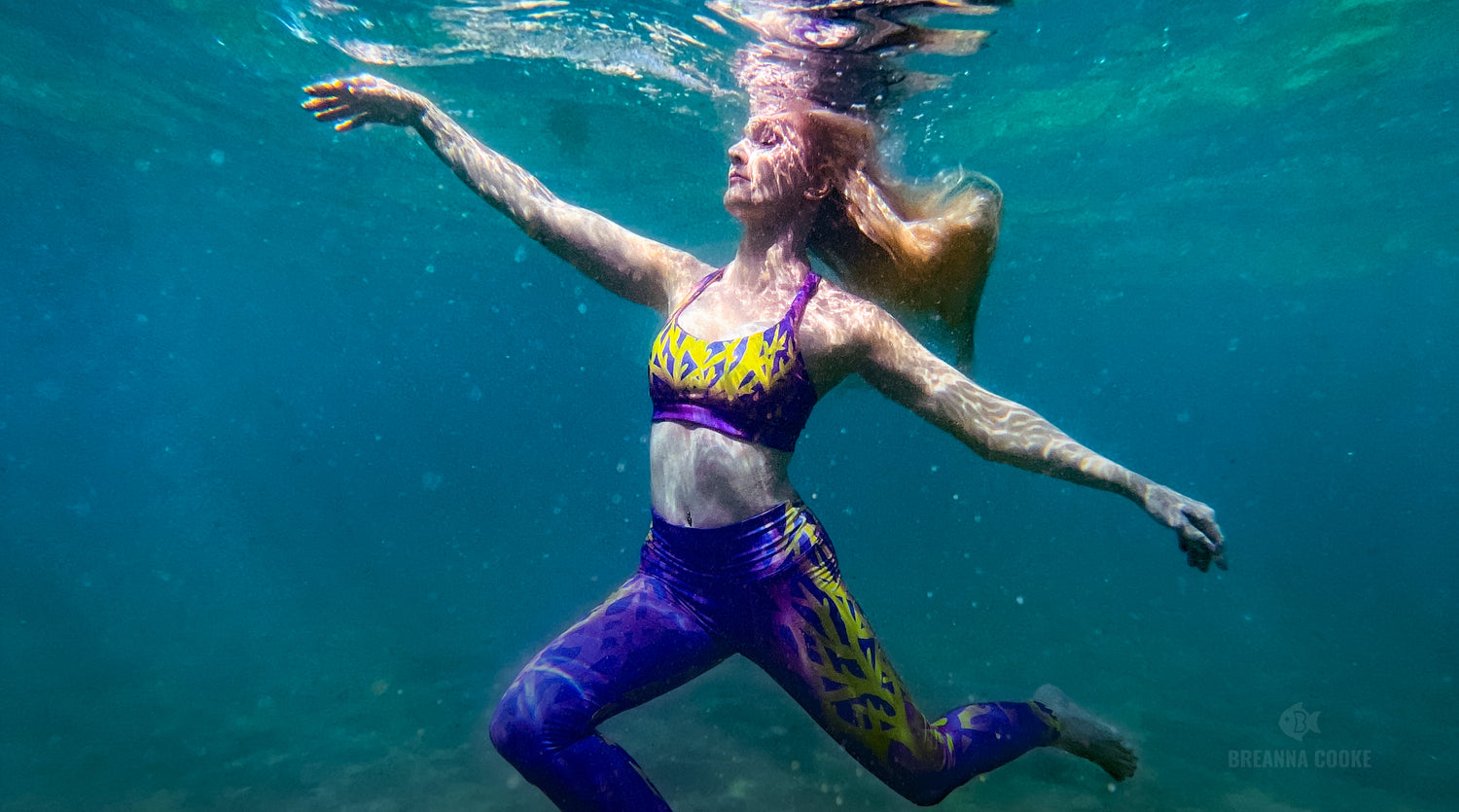 Woman floating underwater in ocean doing graceful dancer pose while wearing purple, yellow, and blue coral reef print leggings and sports bra. Leggings and bra are part of the Glowing Gone climate action campaign. Wear your support for ocean protection.