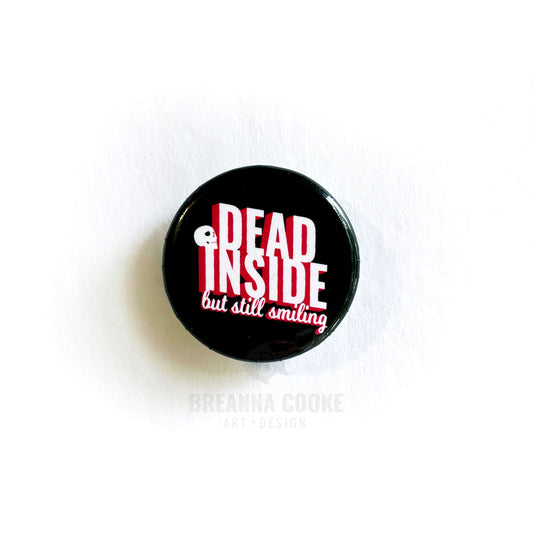 Pin-back button with text "Dead inside but still smiling" on black background.
